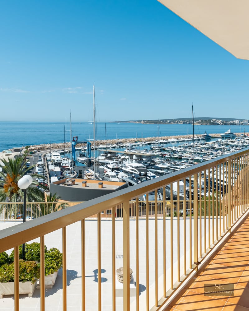 Balcony with a white wicker chair and gold railing overlooking a marina full of boats and the blue Mediterranean Sea.
