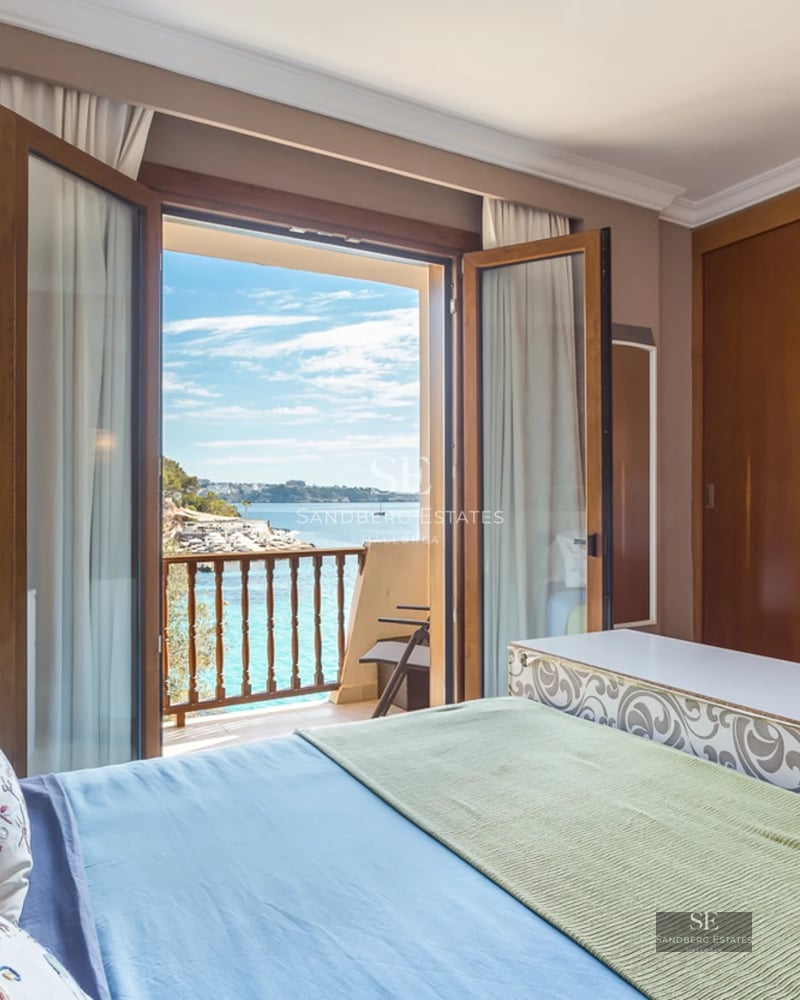 A bedroom with a large bed and open wooden French doors leading to a balcony overlooking the bright blue sea.