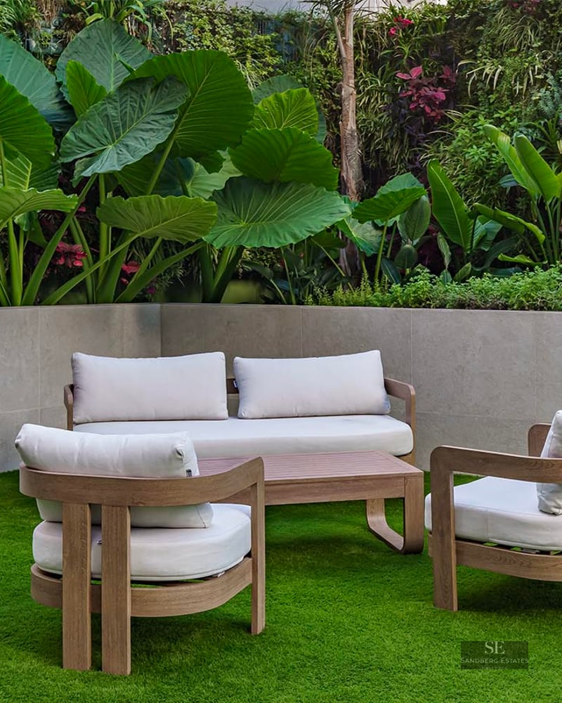 Modern wooden patio furniture with white cushions on green grass next to a vertical garden with tropical plants.