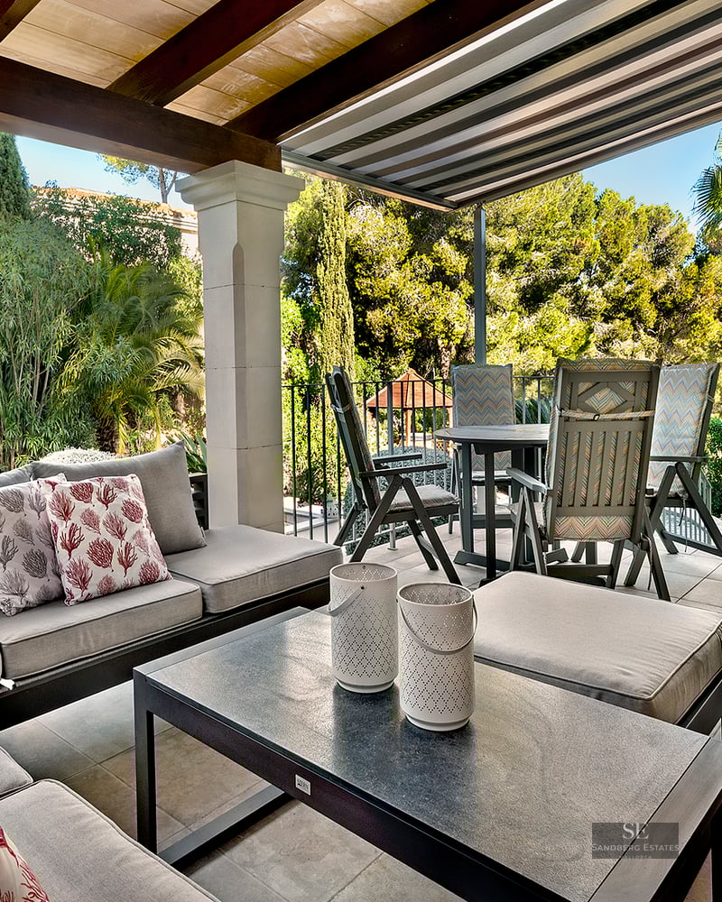 Covered outdoor terrace with lounge sofas, dining table, and view of a lush Mediterranean garden under a bright sky.