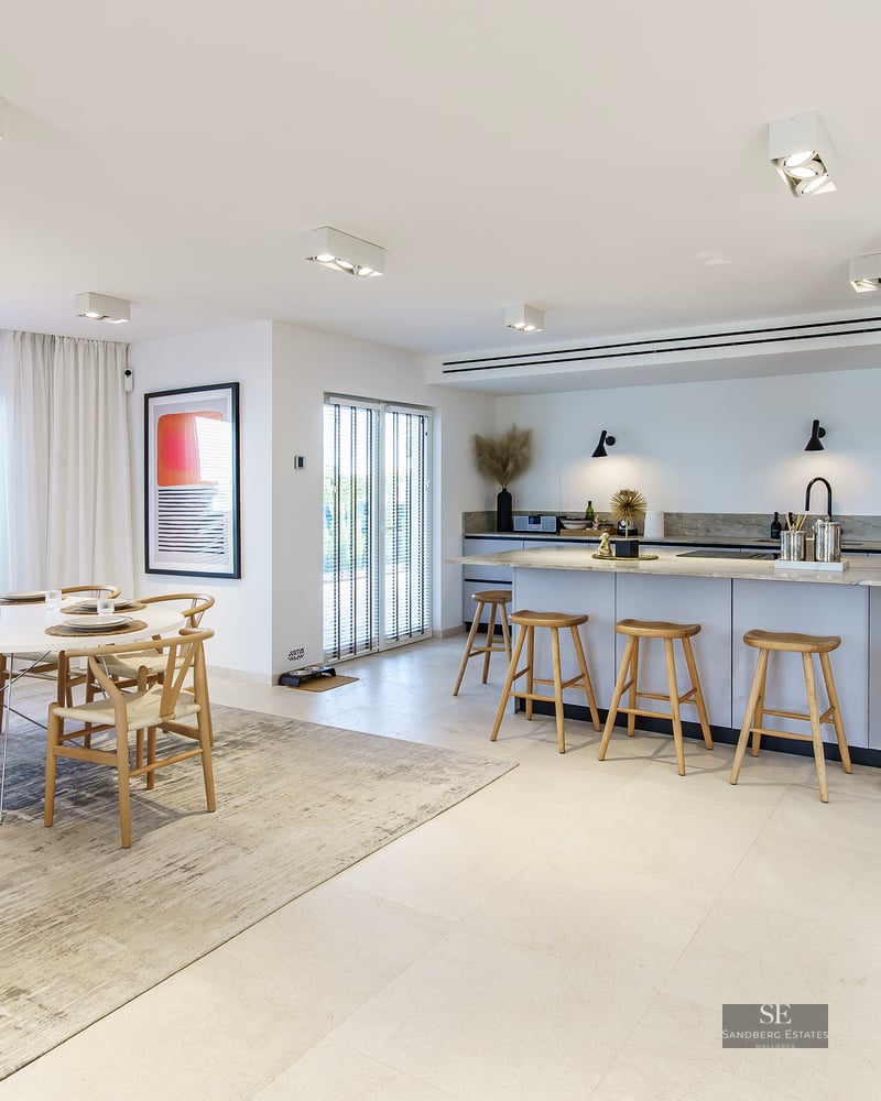 Spacious kitchen featuring a stone island and wooden stools next to a dining area with a round white table and designer chairs.