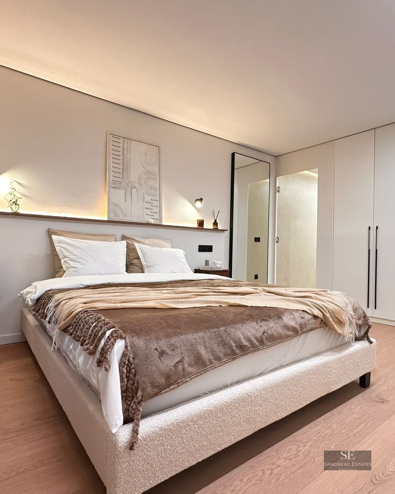 Modern master bedroom featuring a bouclé bed, ambient headboard lighting, light wood floors, and white built-in wardrobes.