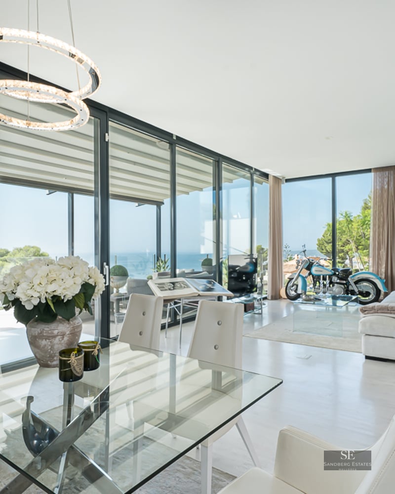 Bright living room with glass dining table, white sofa, floor-to-ceiling windows, and a vintage motorcycle as decor.
