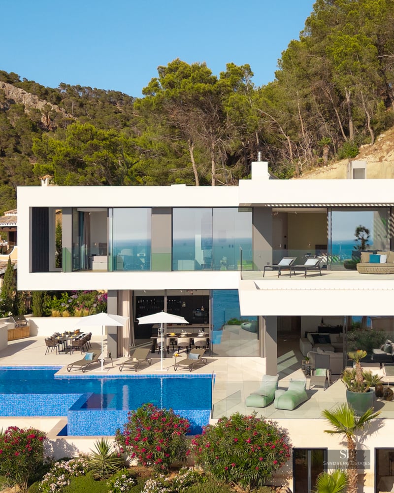 Multi-level modern white villa on a hillside featuring a large infinity pool, lounge terraces, and lush greenery.