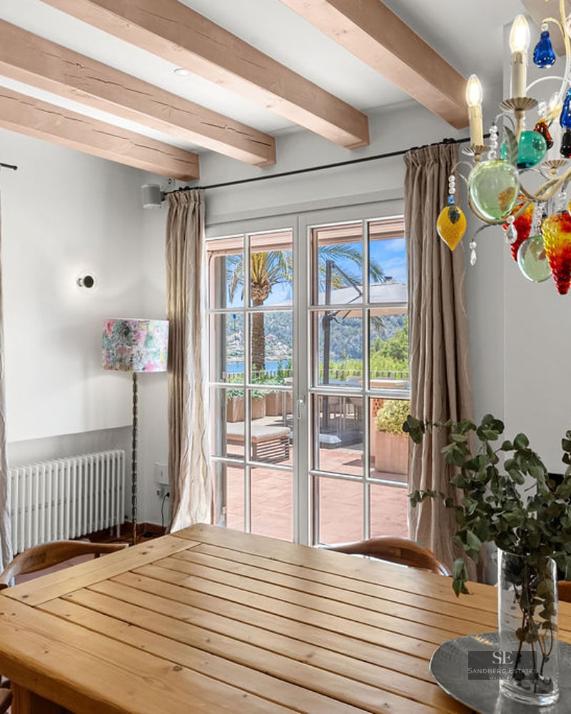 Interior dining room featuring a wooden table, colorful glass chandelier, exposed beams, and doors to a sunny terrace.