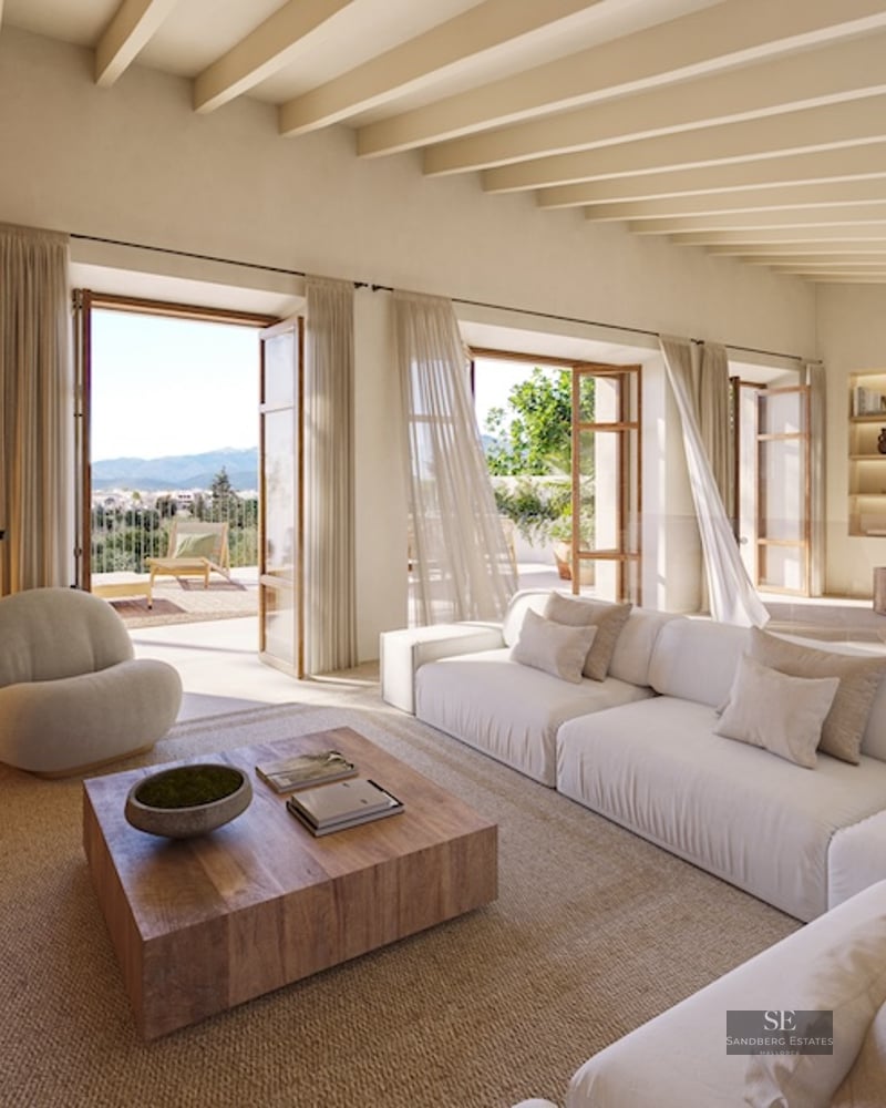 Bright living room with white sofa, stone fireplace, wooden beams, and open doors to a sunny terrace.