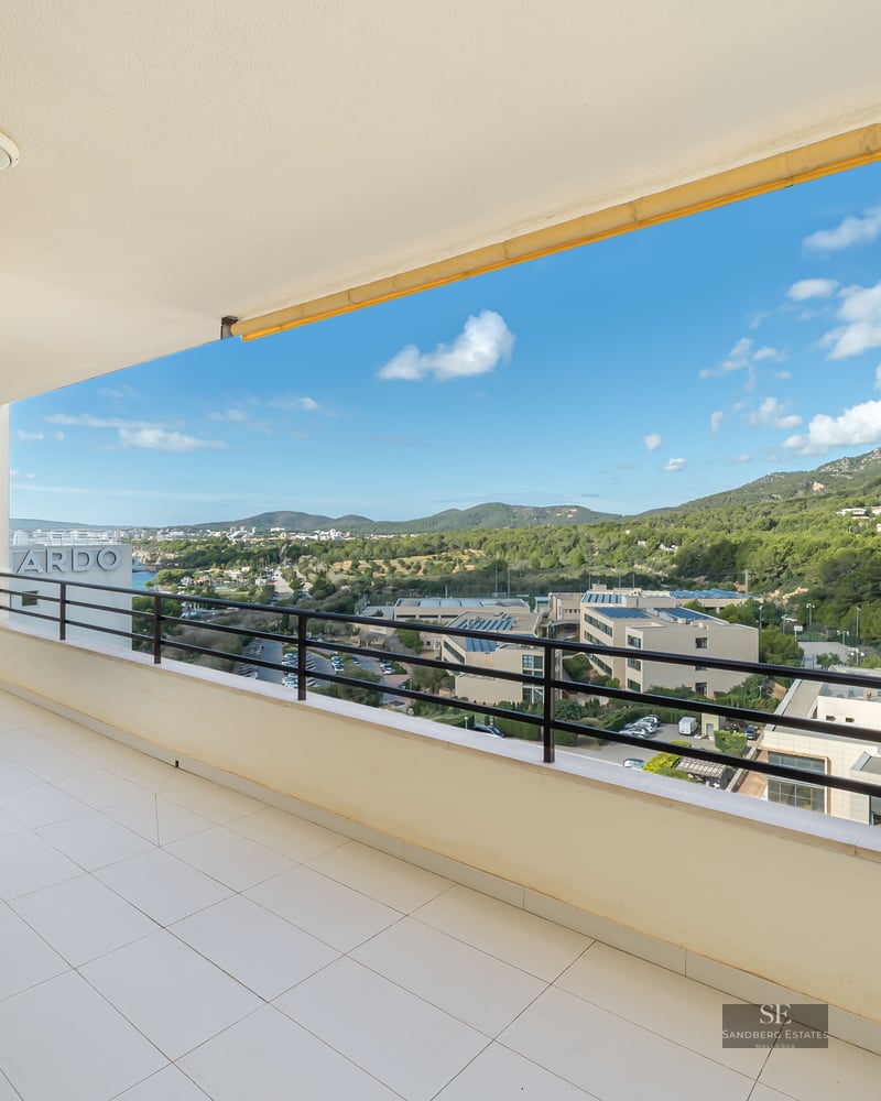 Spacious white tiled balcony with a black metal railing overlooking green hills and a blue sky with white clouds.