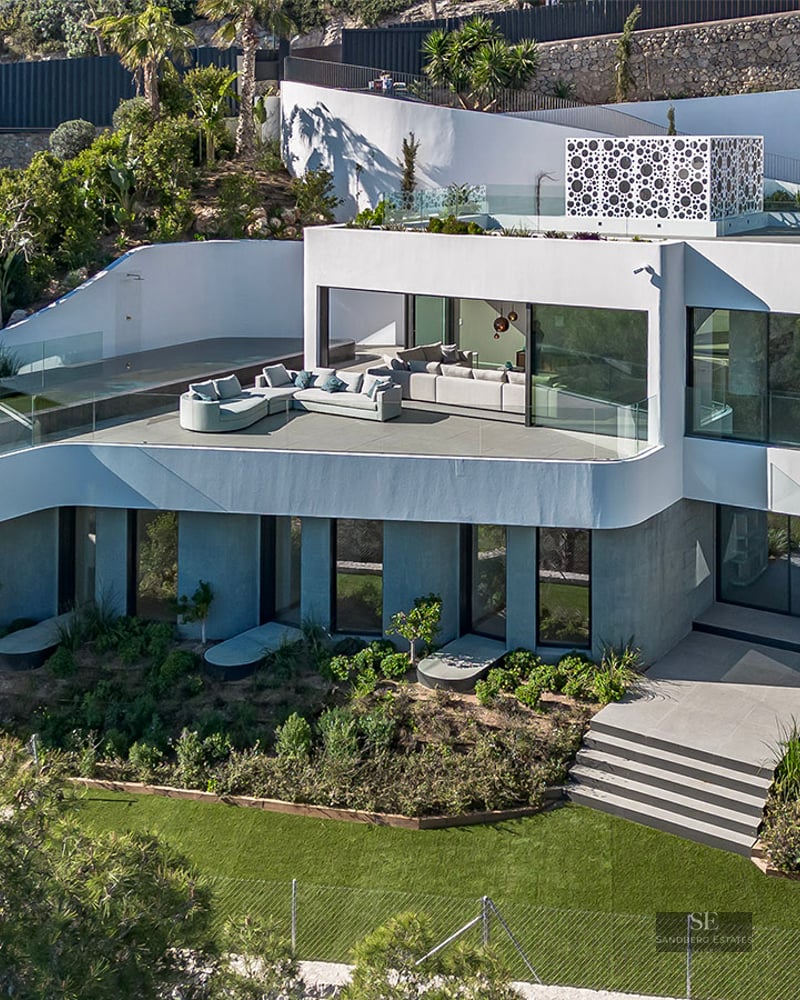 High-angle view of a multi-level white modern villa with an infinity pool, curved terraces, and lush gardens.