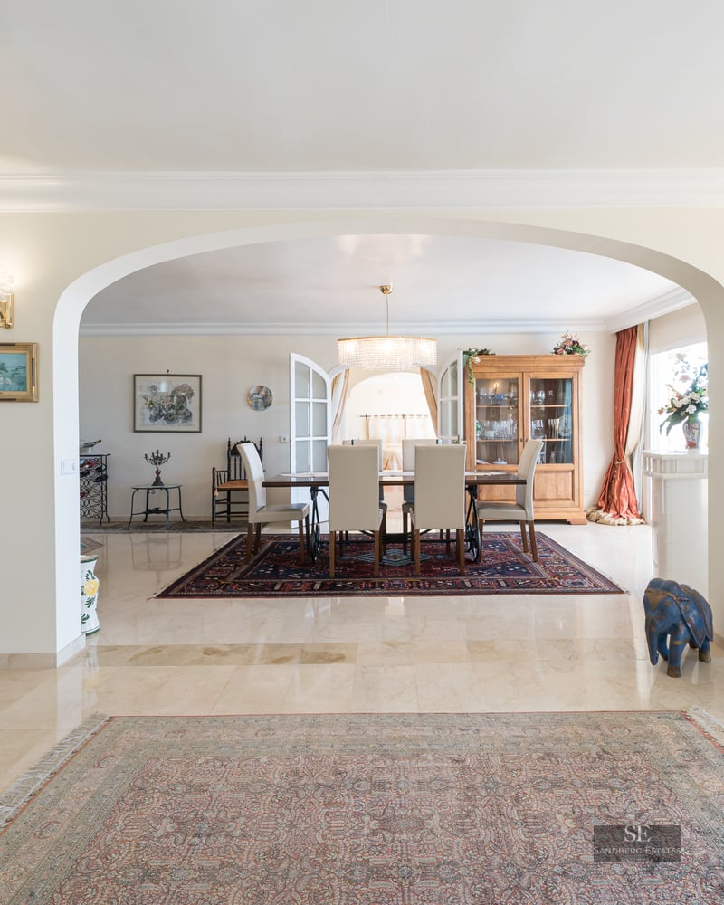 Spacious living area with marble floors and an archway framing a formal dining room with a chandelier and classic rugs.