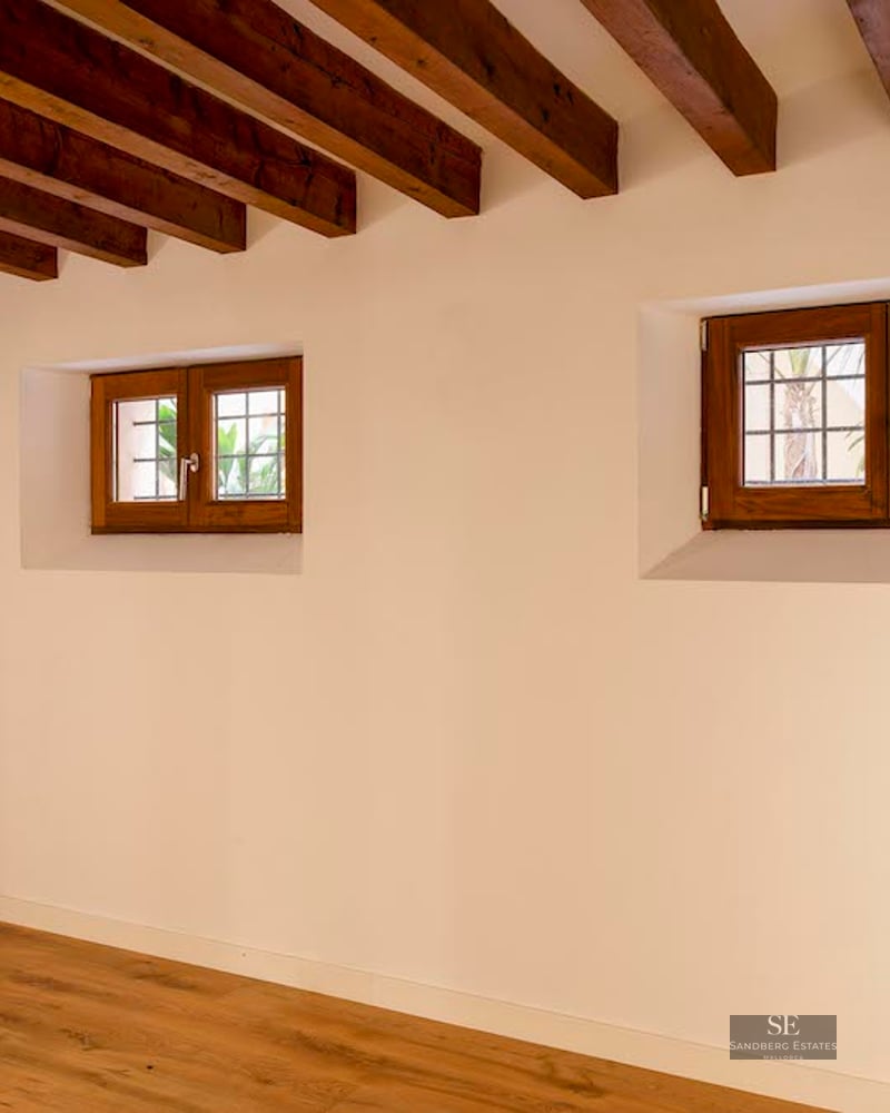 Empty room featuring dark wood-beamed ceiling, white walls, and small wooden windows with hardwood floors.