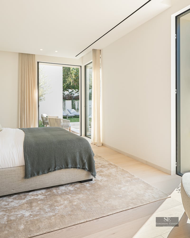 Modern master bedroom with large bed, floor-to-ceiling windows, and views of a lush garden.