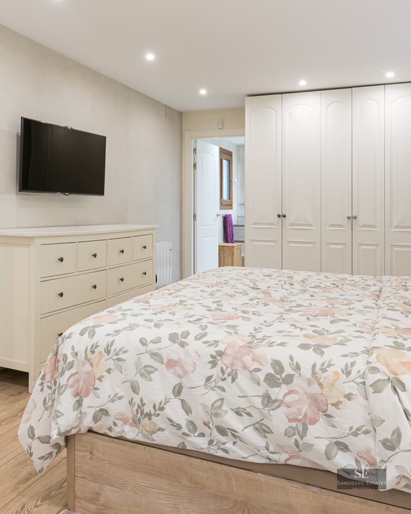 Contemporary bedroom with floral bedding, white dresser, wall-mounted TV, and large floor-to-ceiling wardrobes.