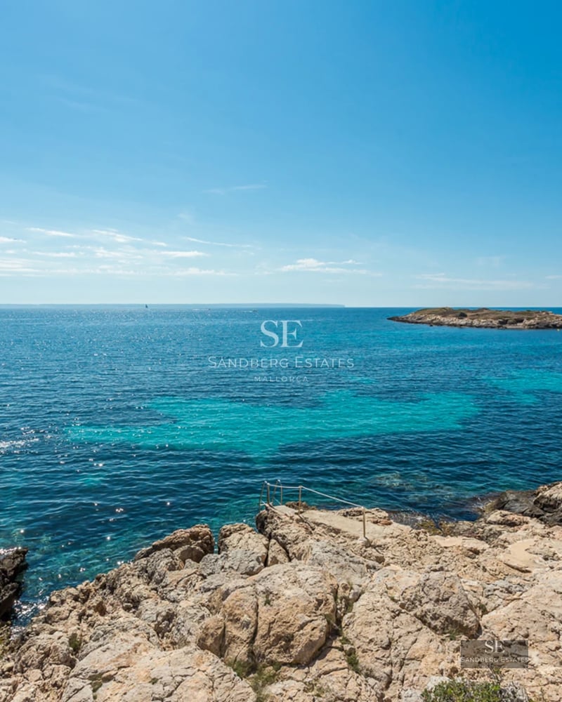 Panoramic view of turquoise Mediterranean waters from a rocky shore with a small island and boat in the distance.