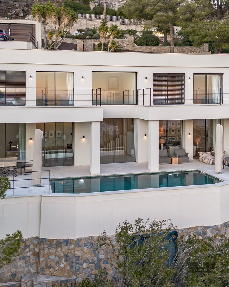 Two-story white modern villa with an infinity pool, spacious terrace, and lush green surroundings on a hillside.