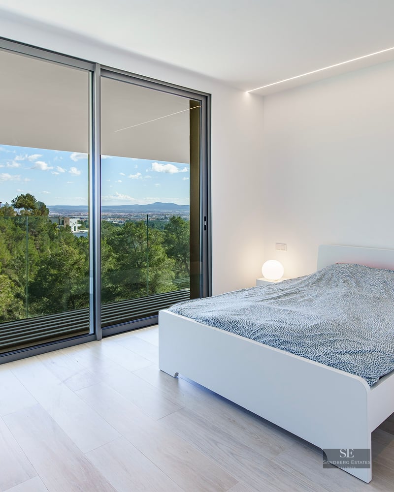 Bright bedroom featuring a white bed, light wood flooring, and large glass doors overlooking a forest and mountains.