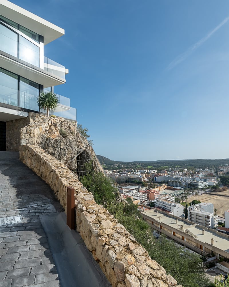 Modern villa with glass walls and stone facade on a cliff overlooking a coastal bay and town under a clear blue sky.