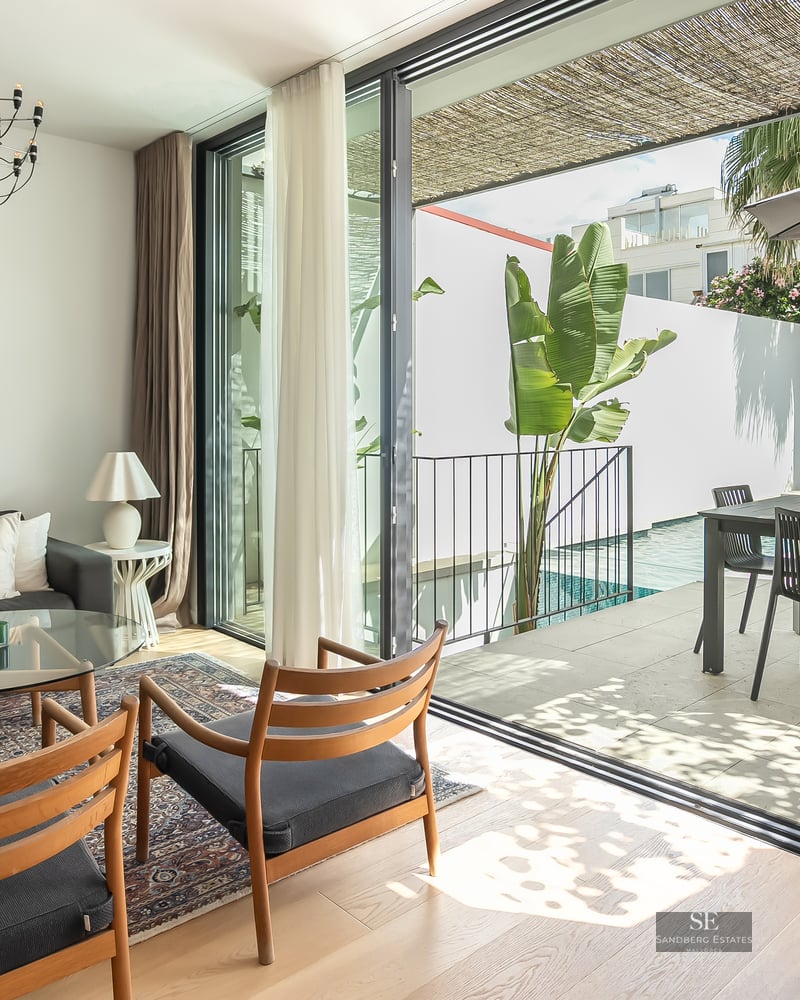 Bright living room with dark sofa and wooden chairs opening to a sunny terrace with a swimming pool and dining area.