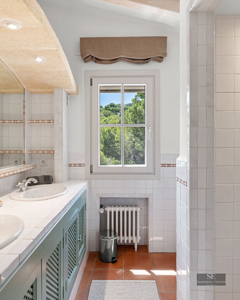 Bright bathroom featuring double white sinks, green cabinetry, terracotta tile floors, and a window viewing lush trees.