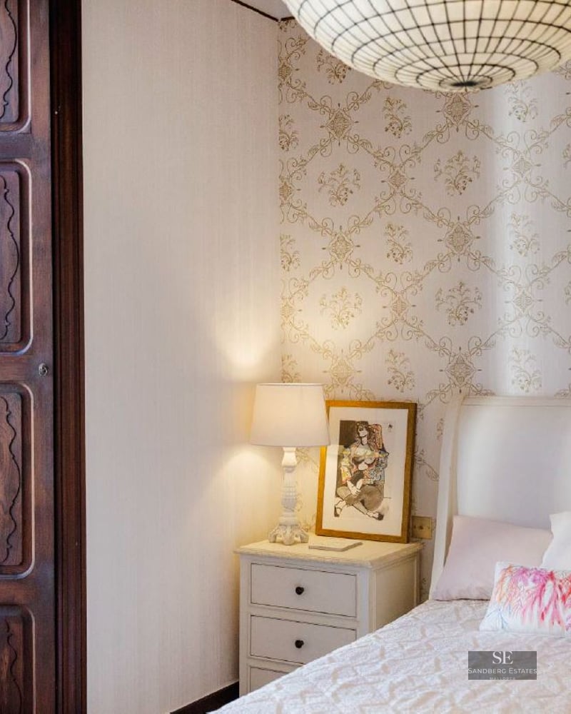 Bedroom featuring patterned wallpaper, a white bed, bedside lamp, and a dark wooden window frame.