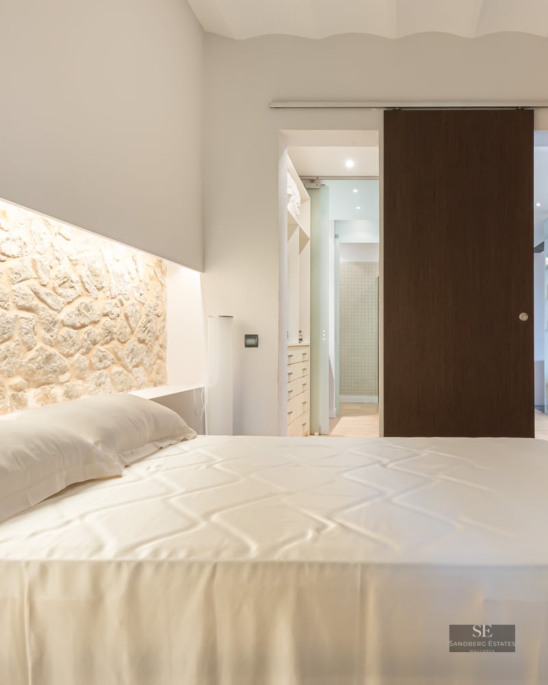 Luxury bedroom featuring a large bed, an illuminated natural stone headboard wall, and a dark wood sliding door.