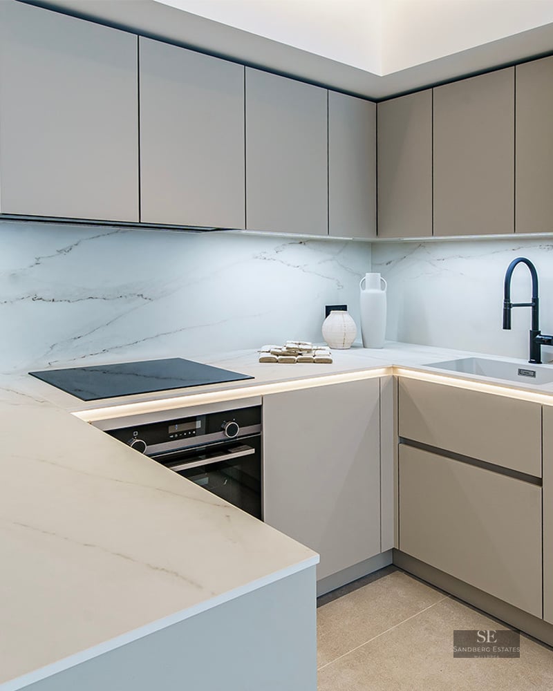 A modern kitchen featuring light gray cabinetry, white marble countertops, LED lighting, and a matte black faucet.