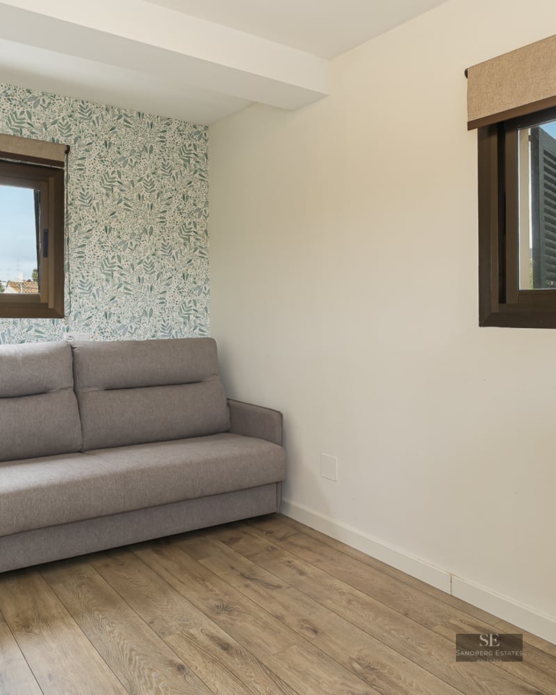 Modern bedroom featuring a grey sofa, botanical leaf pattern wallpaper, wooden floors, and natural light from two windows.
