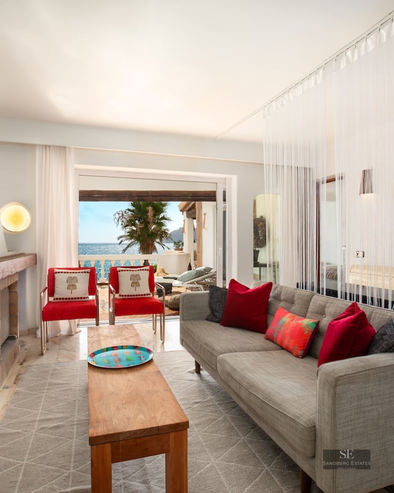 Living room featuring a grey sofa, red chairs, rustic fireplace, and large windows overlooking the sea and terrace.