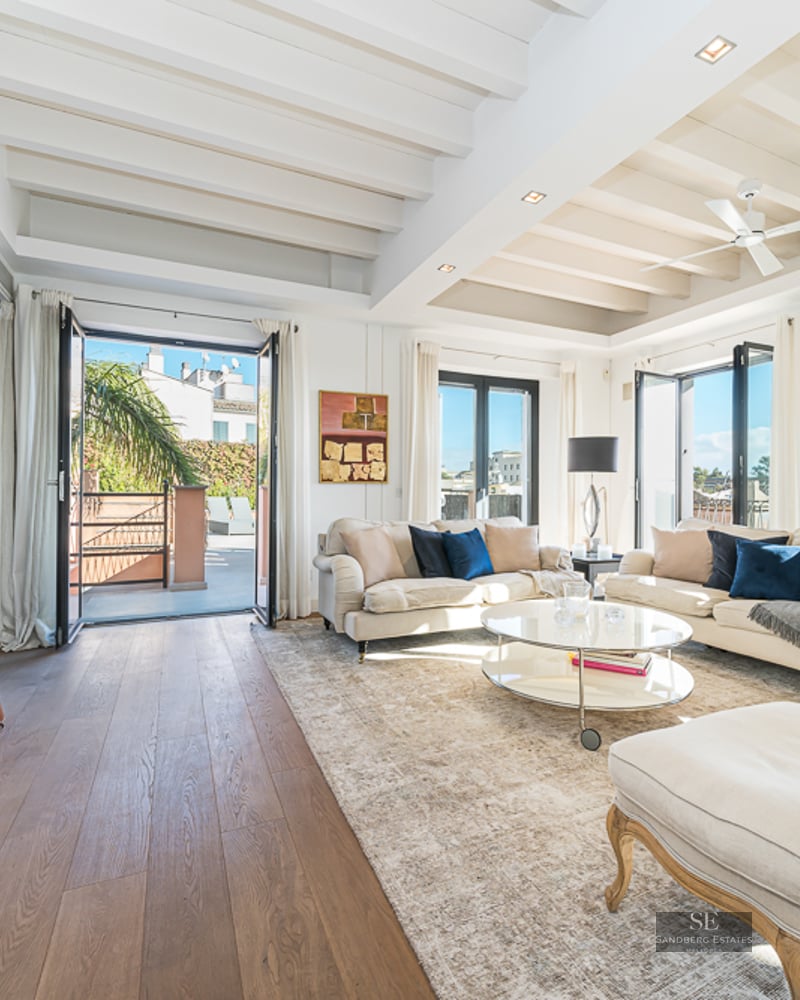Elegant living room featuring cream sofas, white beamed ceilings, and access to a sunny outdoor terrace.
