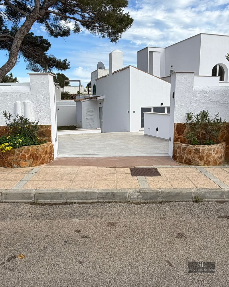 Entrance to a white villa featuring stone-clad walls, planters with yellow flowers, and a paved driveway under pine trees.