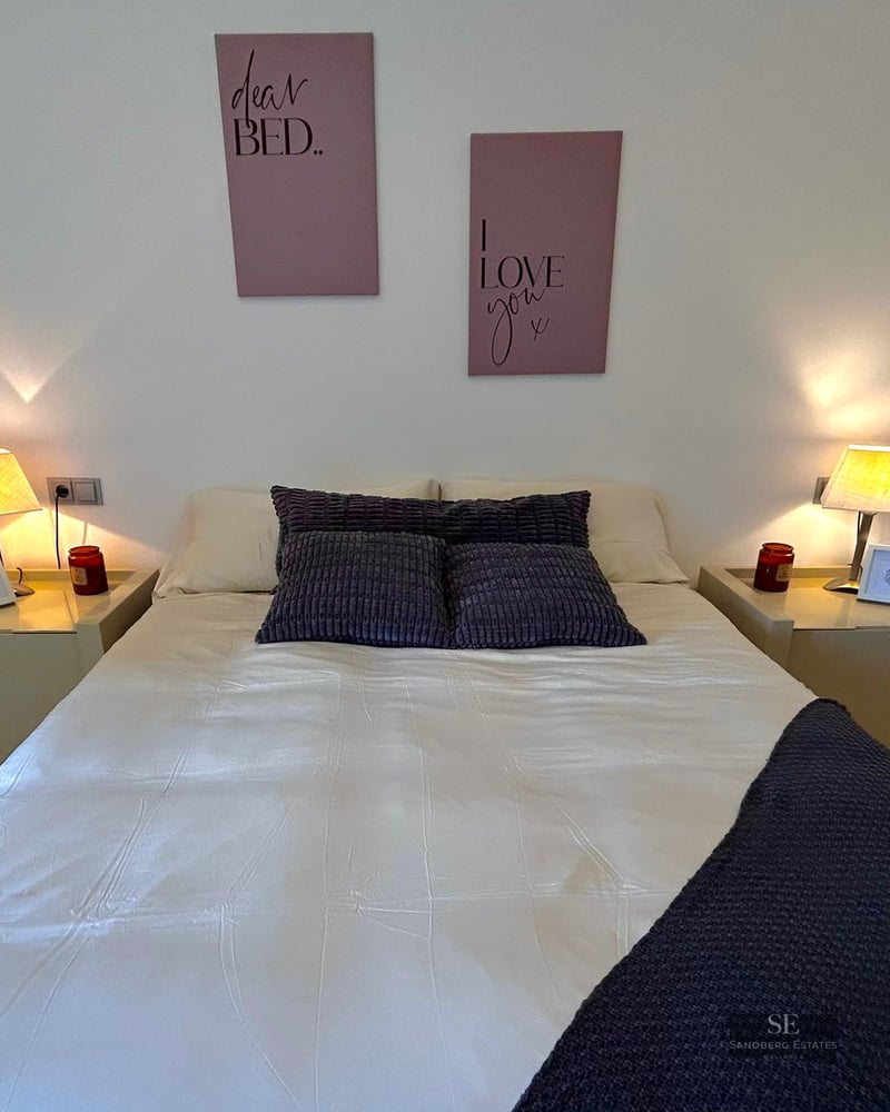 Neatly made bed with white linens and purple pillows between two glowing bedside lamps in a modern room.