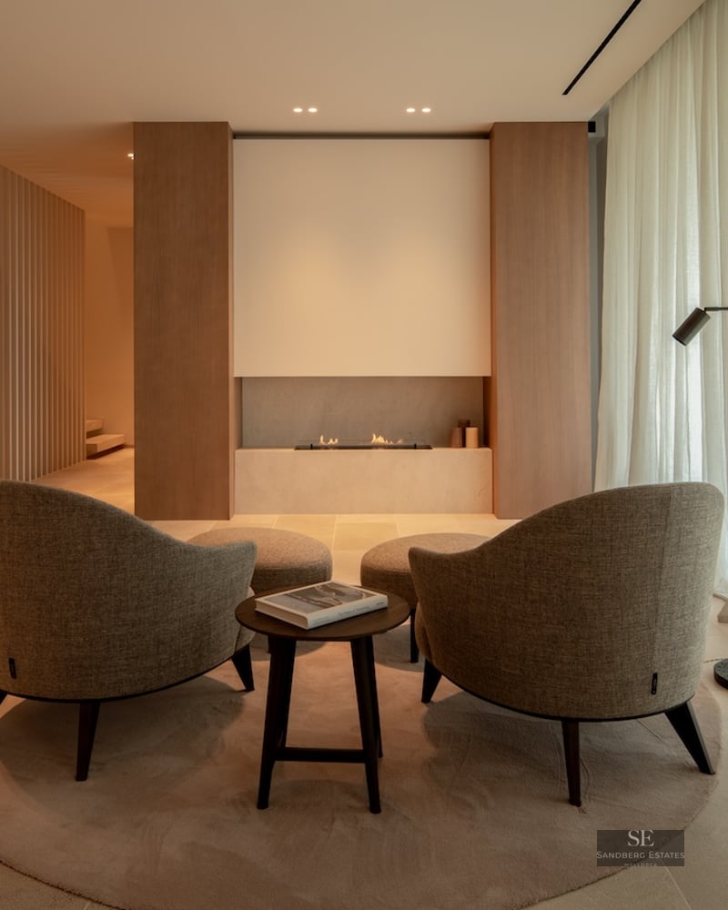 Minimalist lounge featuring two beige armchairs facing a built-in linear fireplace, warm ambient lighting, and wood paneling.