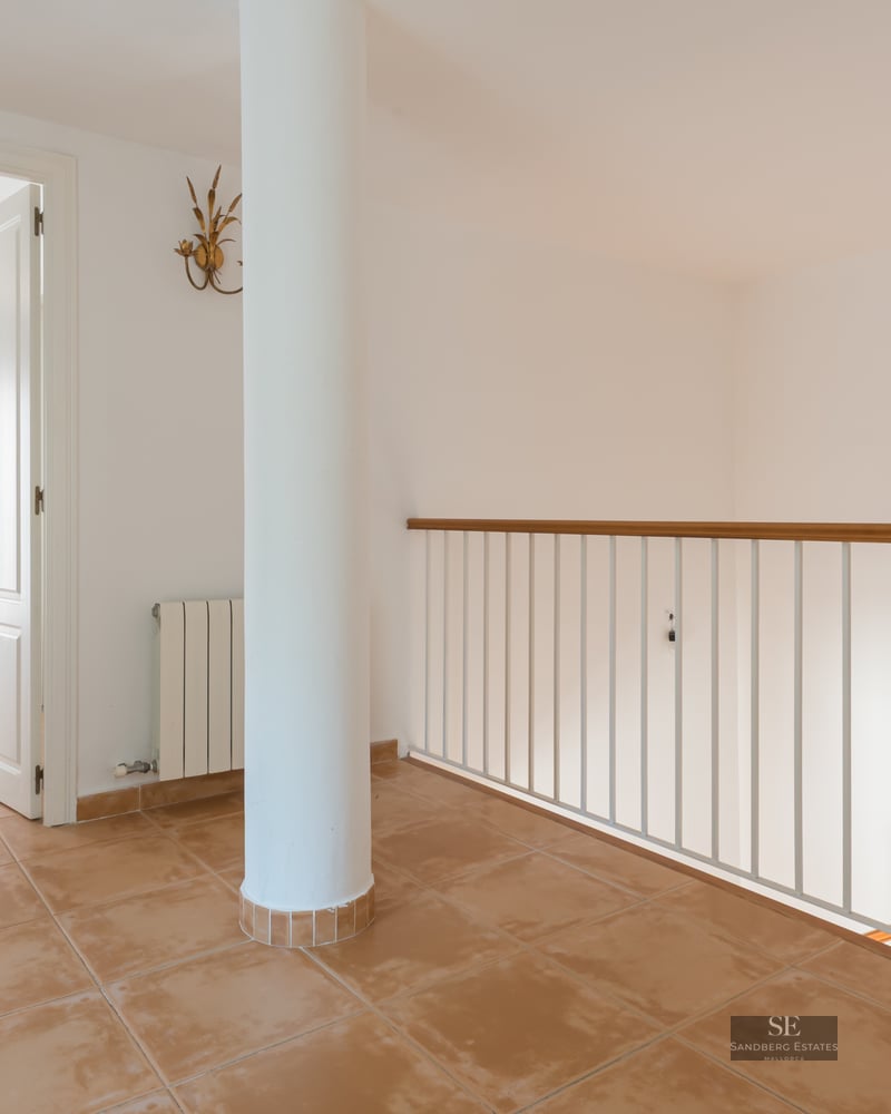 Hallway with terracotta tiles, a white pillar, and a staircase railing, leading to an open doorway.