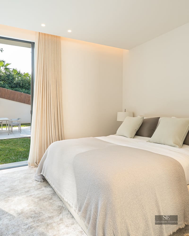 A bright, modern bedroom with a large bed opening onto a sunny private garden through glass sliding doors.
