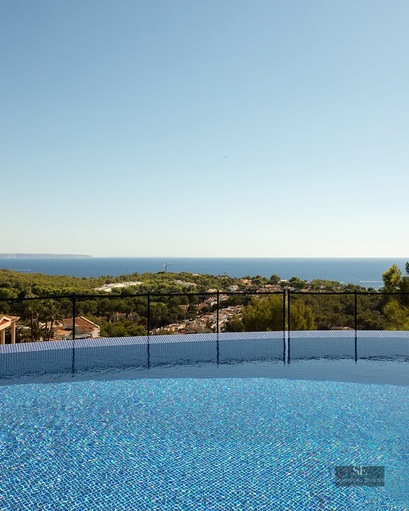 View from a blue mosaic infinity pool looking out over green trees towards the Mediterranean sea under a clear sky.