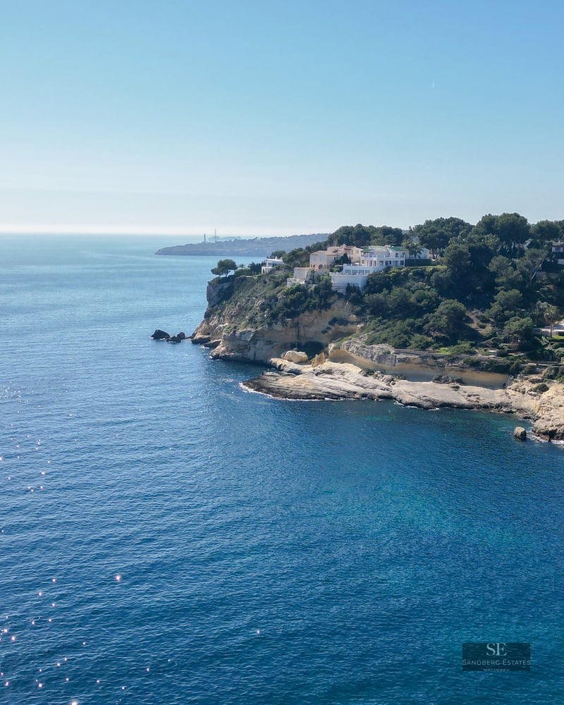 Drone shot of rocky cliffs with luxury white villas overlooking the sparkling blue Mediterranean sea.
