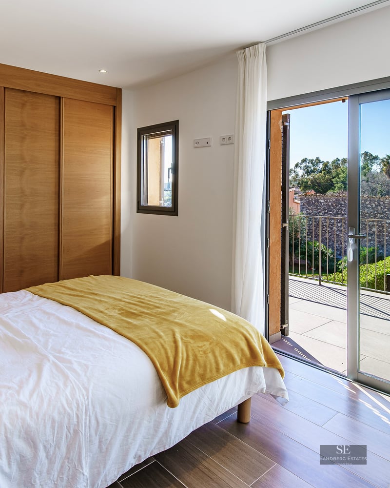 Bright bedroom with wooden built-in closets and large sliding glass doors opening onto a sunny terrace with green views.