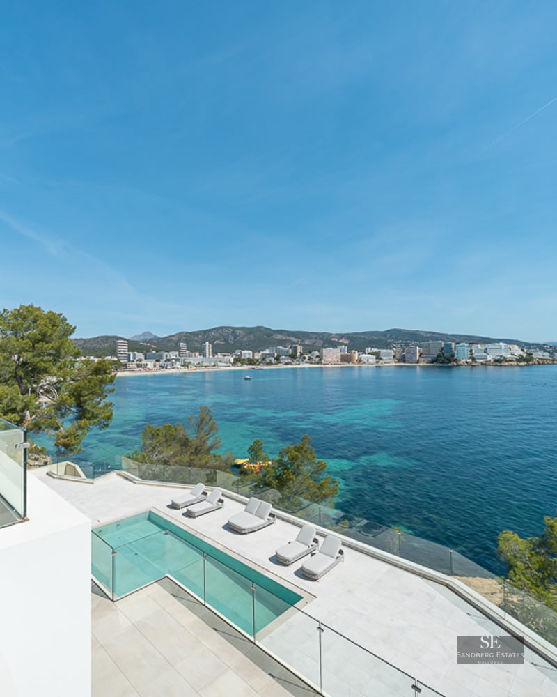 Modern white terrace with infinity pool and glass railings overlooking a turquoise bay and coastline.