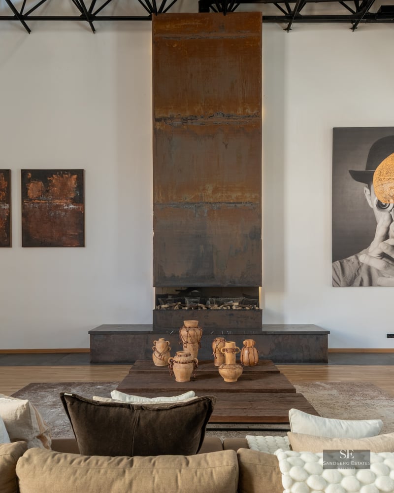 Spacious living room with a tall weathered steel fireplace, beige sofa, and woven pendant lamps in an industrial loft.