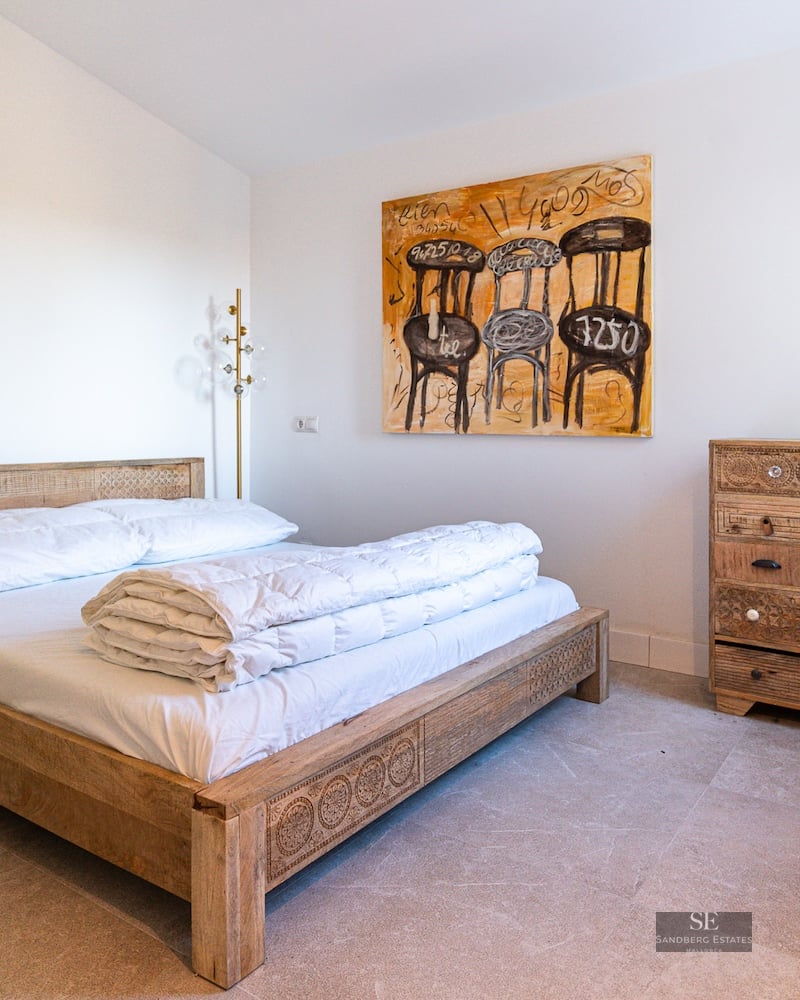 Bright bedroom with a carved wooden bed, matching dresser, large abstract chair painting, and natural stone floors.