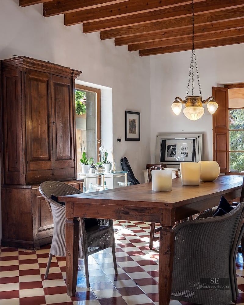 A rustic dining room with a large wood table, red and white checkered tile floor, and exposed ceiling beams.