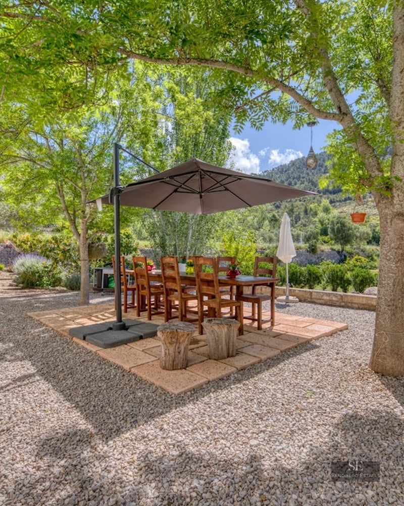 Wooden dining table and chairs under a parasol on a gravel terrace surrounded by trees and mountains.
