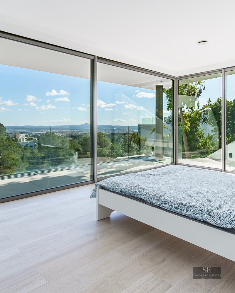 Modern bedroom with white bed and large floor-to-ceiling glass walls overlooking a lush green forest and distant city.