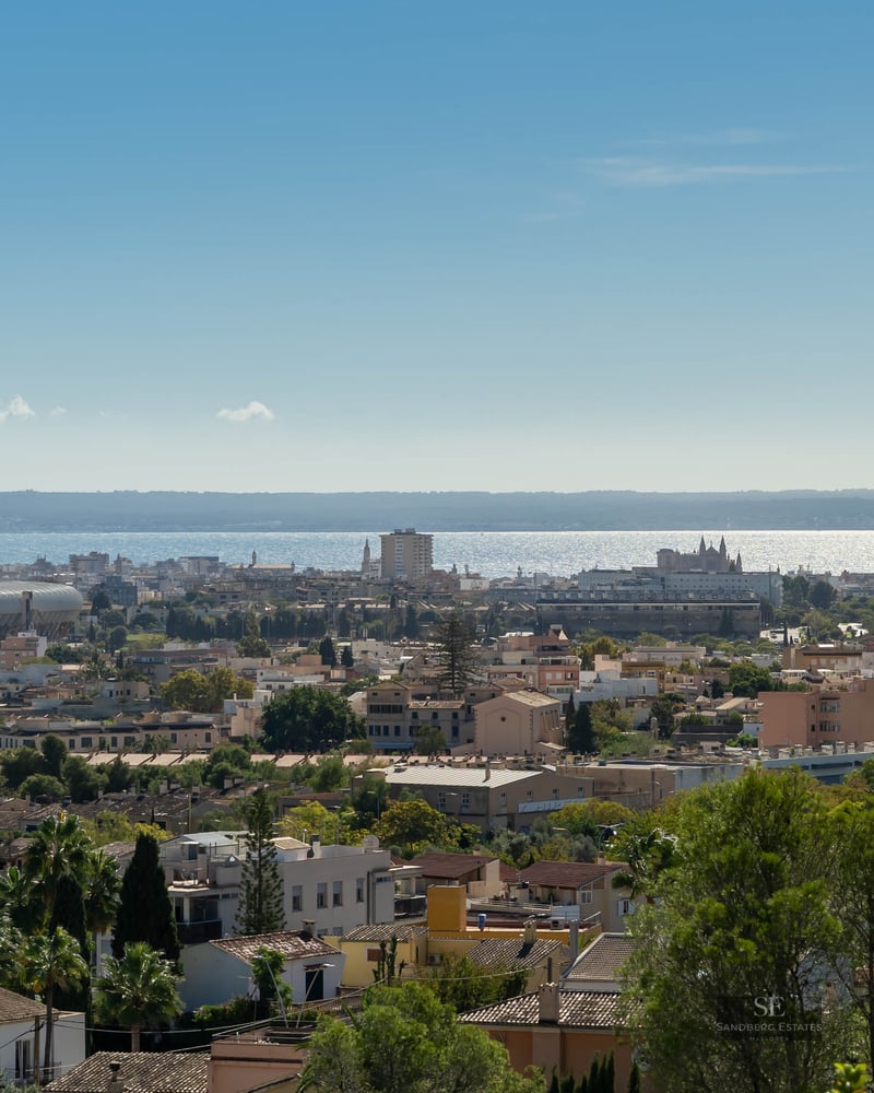 Wide panoramic view of Palma with buildings, greenery, and the shimmering Mediterranean Sea on the horizon.