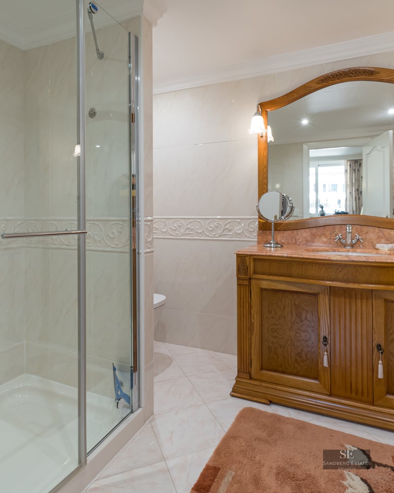 A classic bathroom featuring a glass-enclosed shower, cream marble wall tiles, and a large wooden vanity with a mirror.