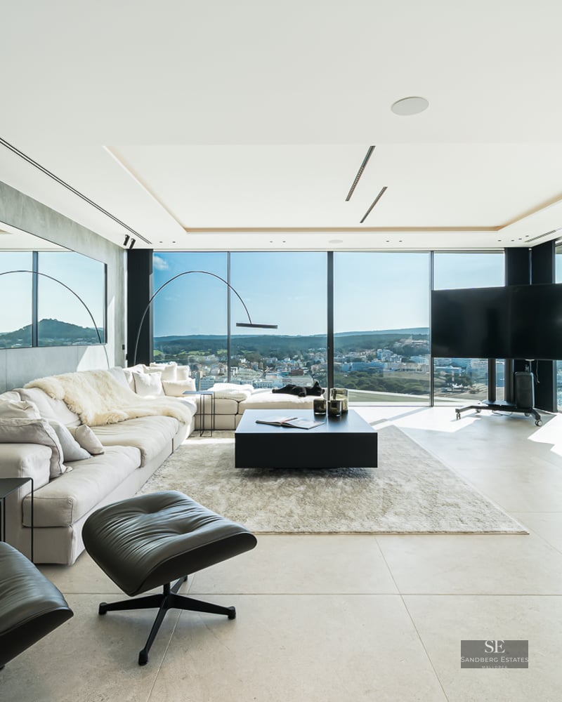 Modern living room with a large white sofa, black leather lounge chair, and floor-to-ceiling windows showing a coastal view.