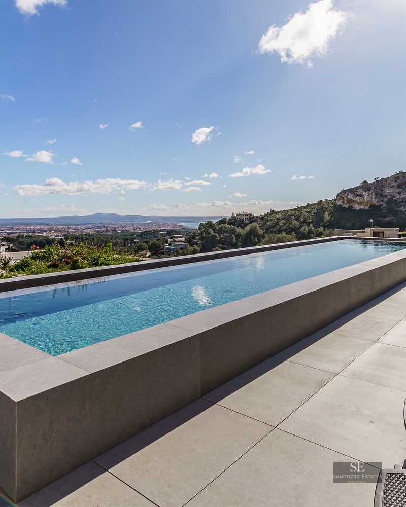 Infinity pool on a stone terrace with sun loungers overlooking a valley and city landscape.