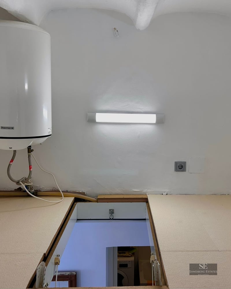 White utility room with an electric water heater on the wall and a rectangular hatch open to the floor below.