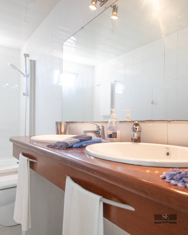 Modern white bathroom featuring a wooden double sink vanity, large mirror, and bathtub with glass screen.