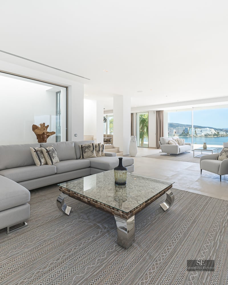 Spacious modern living room with light gray sofas, a glass coffee table, and large windows overlooking the Mediterranean sea.