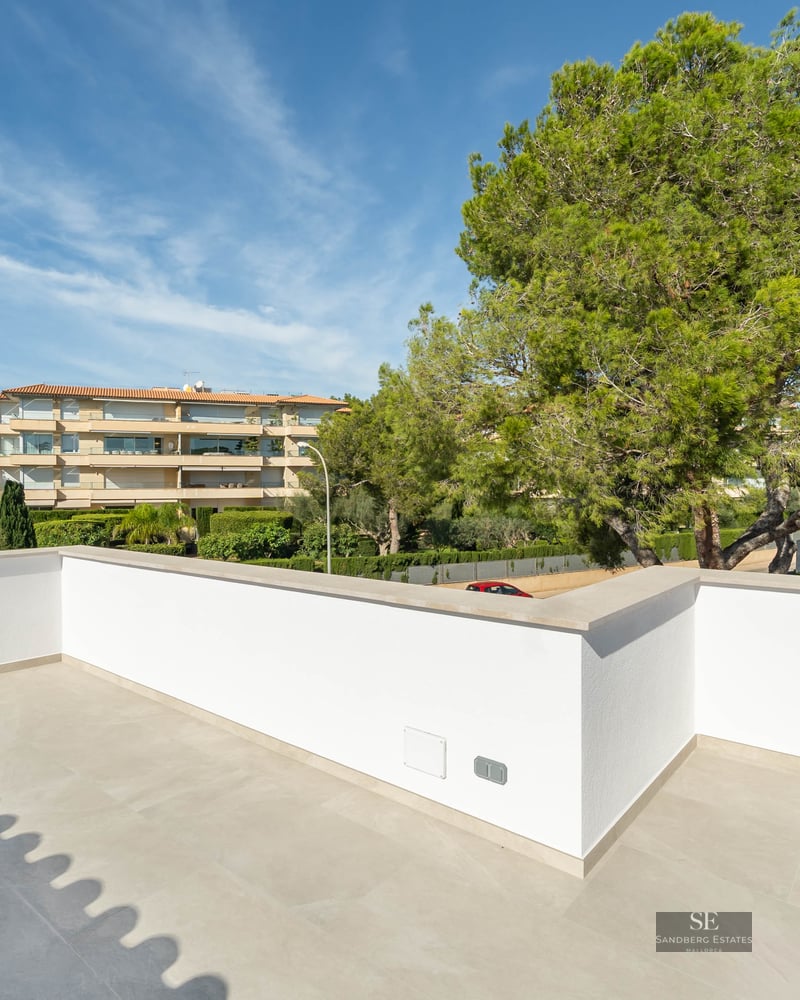 Spacious modern terrace with white walls and light stone flooring, overlooking lush pine trees and buildings.